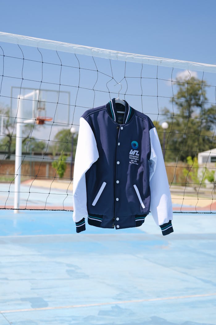 A navy and white varsity jacket hangs on a volleyball net in a sunny outdoor sports court.